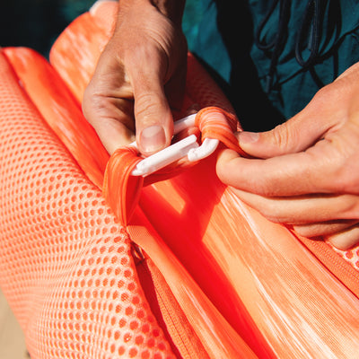 Close-up of hands adjusting a white strap on an orange mesh hammock. 