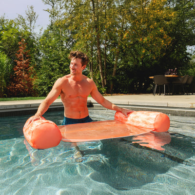 Man holding an pool float in a swimming pool with trees and a table in the background. 