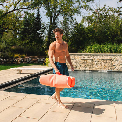 Man holding a pink pool noodle by a pool with trees in the background 