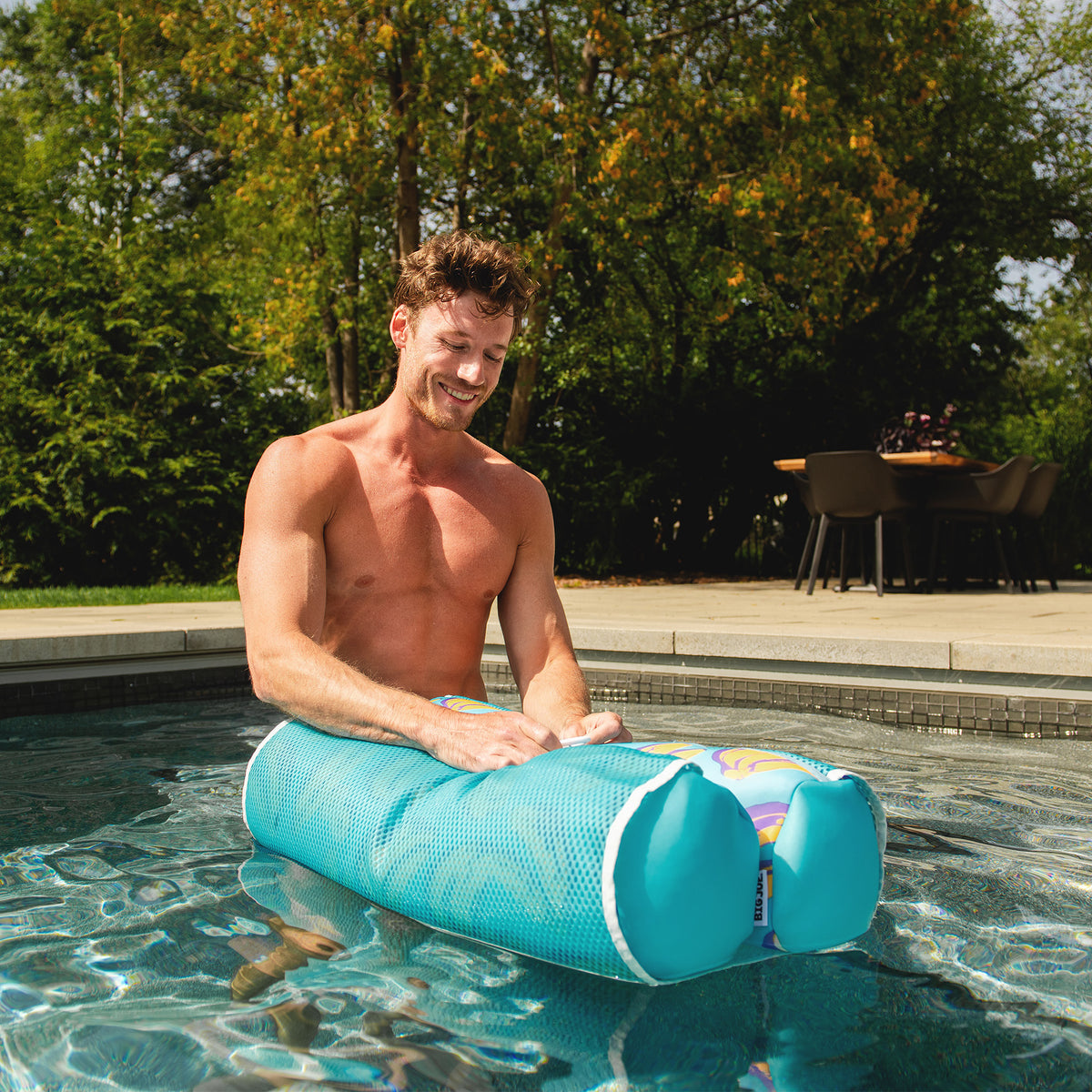Man using a blue pool noodle in a swimming pool with trees and outdoor furniture in the background 