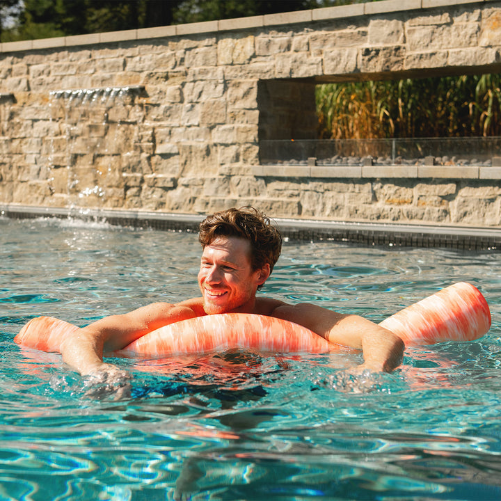 Man floating on a pink pool noodle in a pool with a stone wall and plants in the background #color_peach-glow