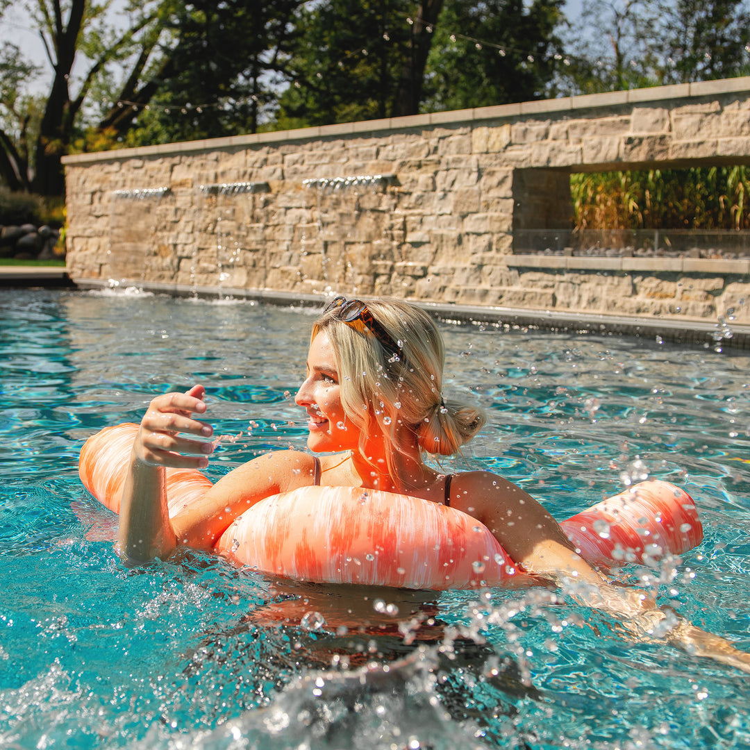 Woman floating on a pink flamingo pool float in a swimming pool with a stone wall and trees in the background. #color_peach-glow