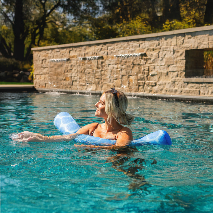 Woman relaxing in a pool with pool noodle floats, surrounded by a stone wall and greenery. #color_coral-bloom