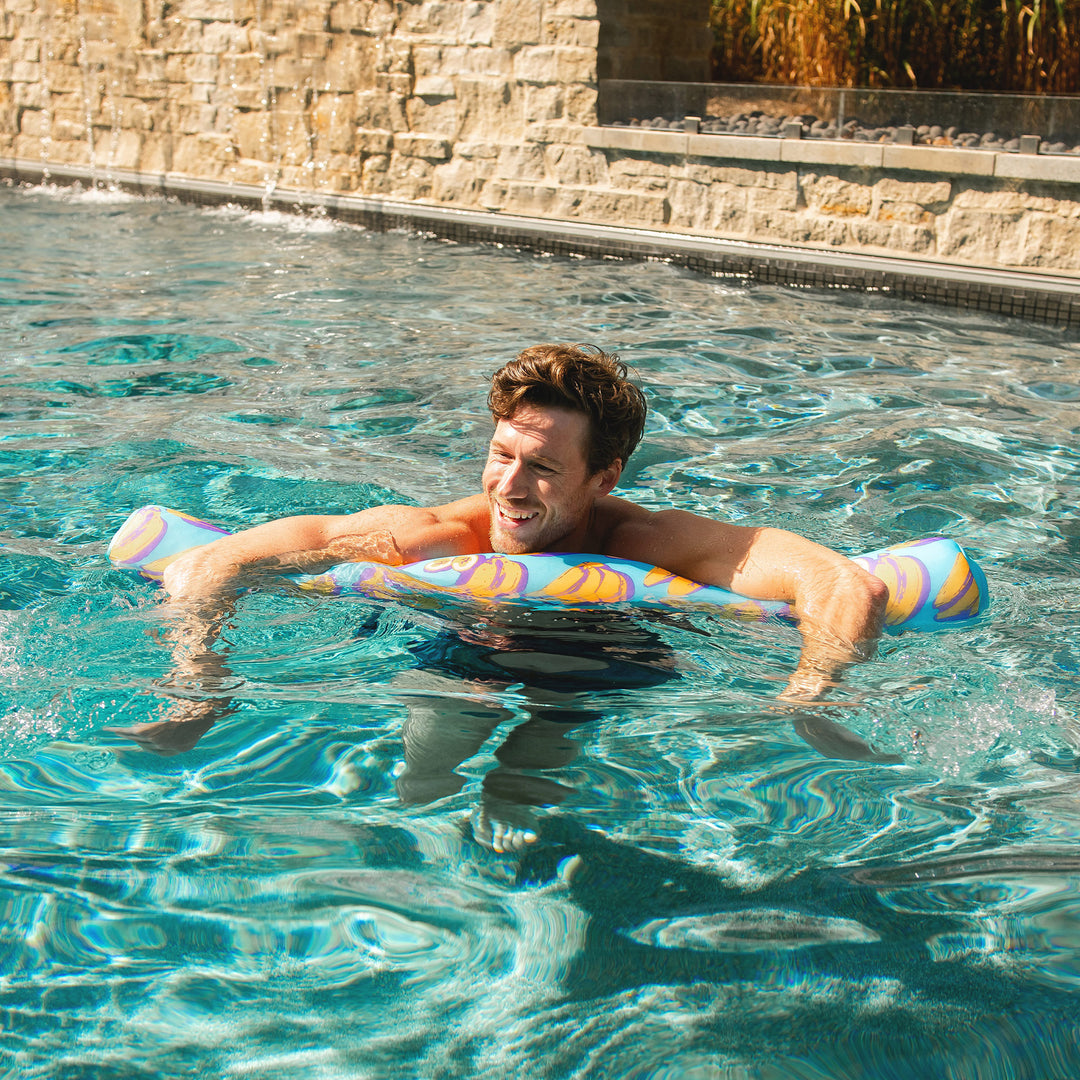 Man floating on a colorful pool noodle in a pool with stone wall and plants in the background #color_banana-pop