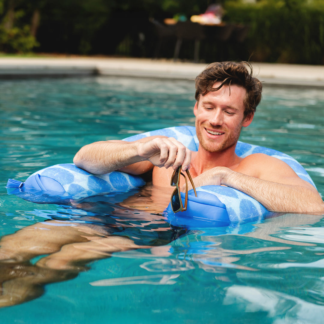 Man floating on a blue noodle sling in a pool, holding sunglasses. #color_coral-bloom