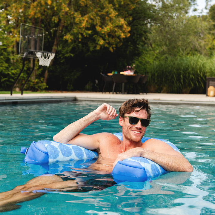 Man lounging on a blue pool float in a pool with trees and outdoor furniture in the background. #color_coral-bloom