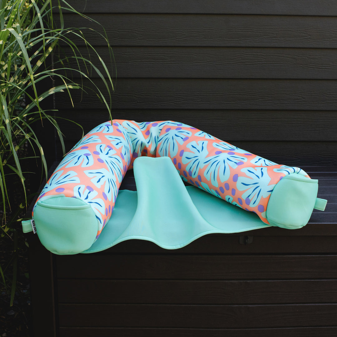 Multicolored U-shaped pool float chair on a green mat against a dark wooden  #color_sweet-shells-peachwall.