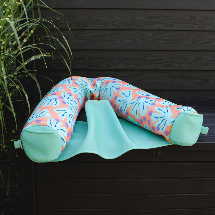 Multicolored U-shaped pool float chair on a green mat against a dark wooden  #color_sweet-shells-peachwall.