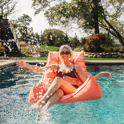 Woman lounging on a pink chair in a pool with trees and garden in the background 