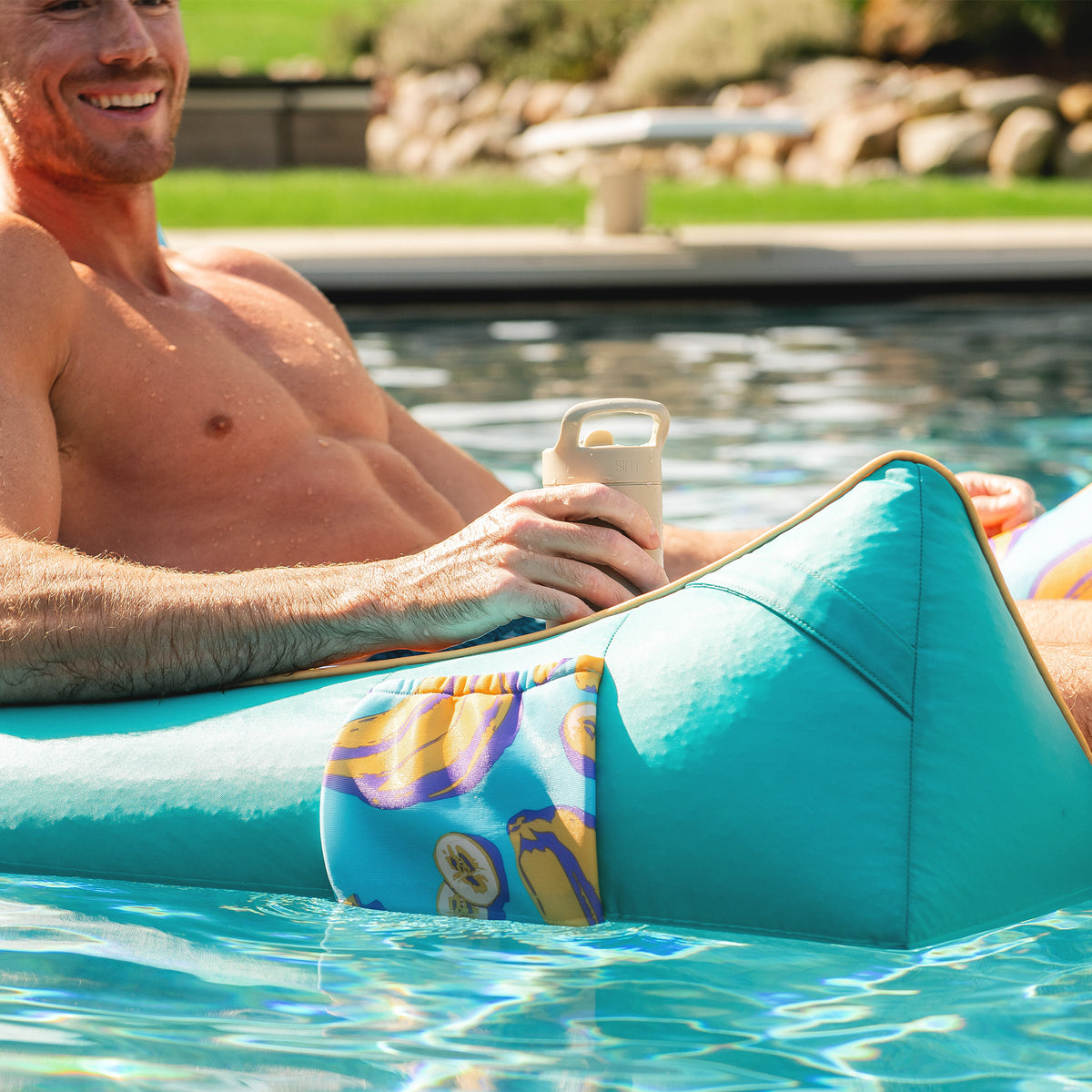 Man lounging on a teal pool float in a pool, holding a drink. 