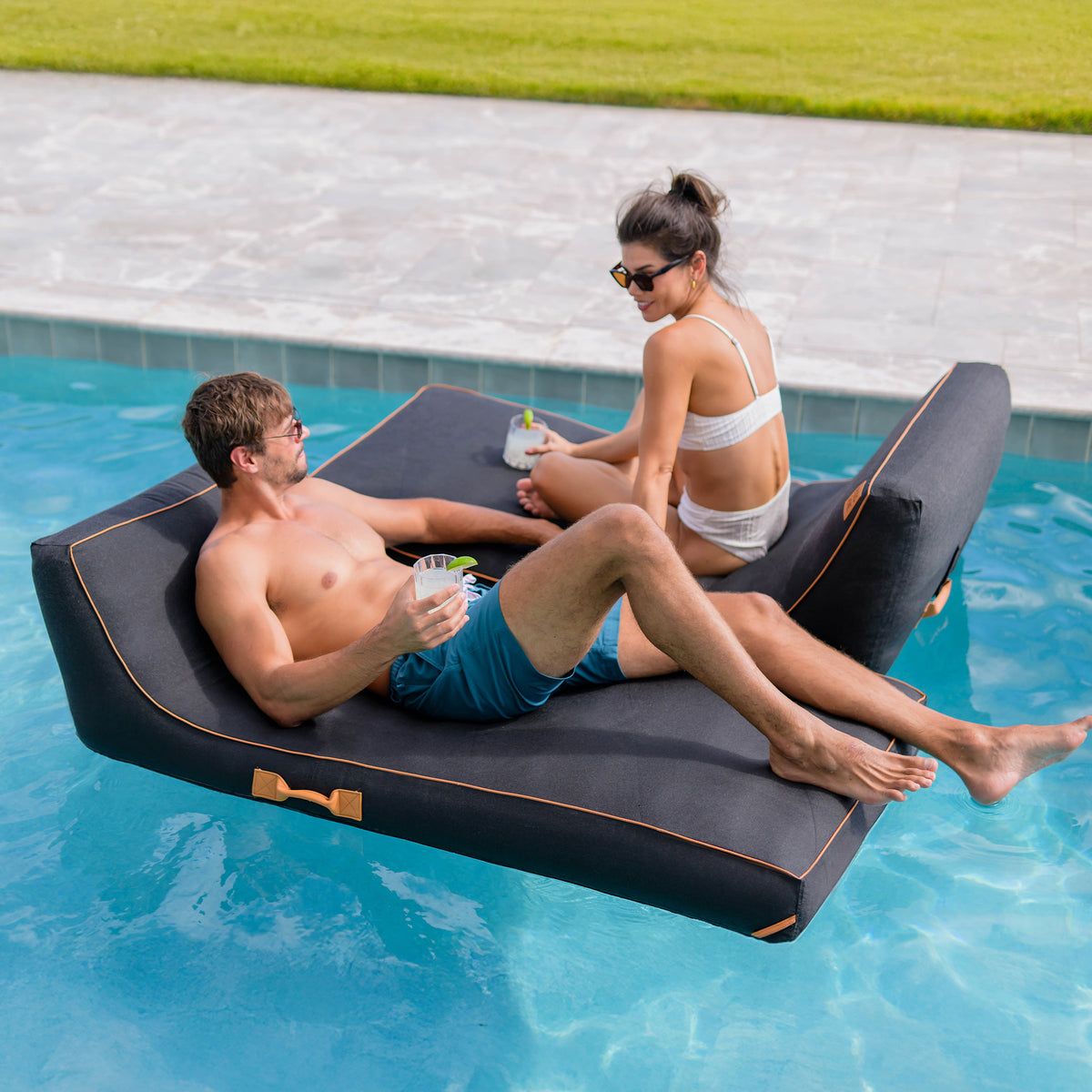 Two people relaxing on a floating inflatable lounge in a pool. #color_sand-stripe-sunbrella-fabric