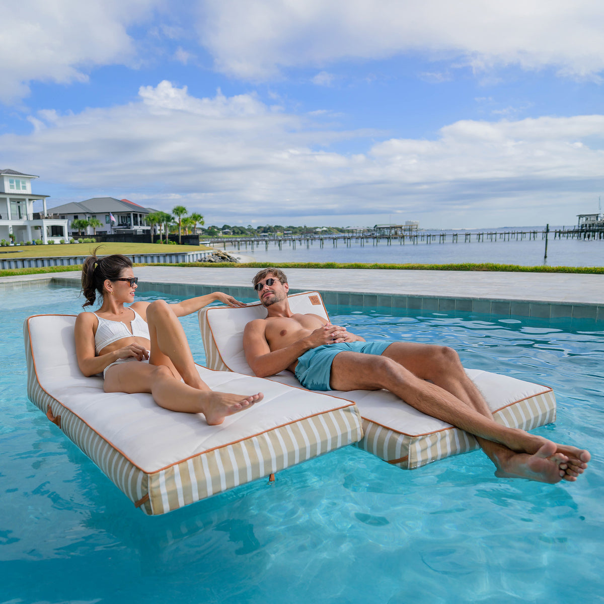 Two people relaxing on a floating platform in a pool with a scenic background. 