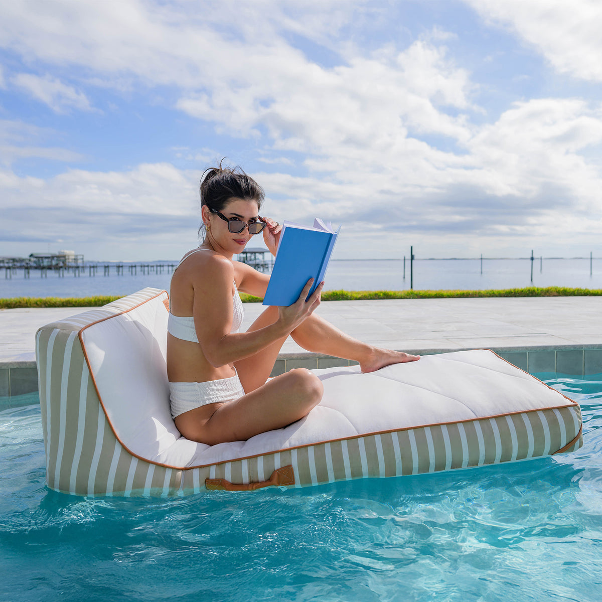 Woman reading a book on a striped pool float in a pool with a scenic background 