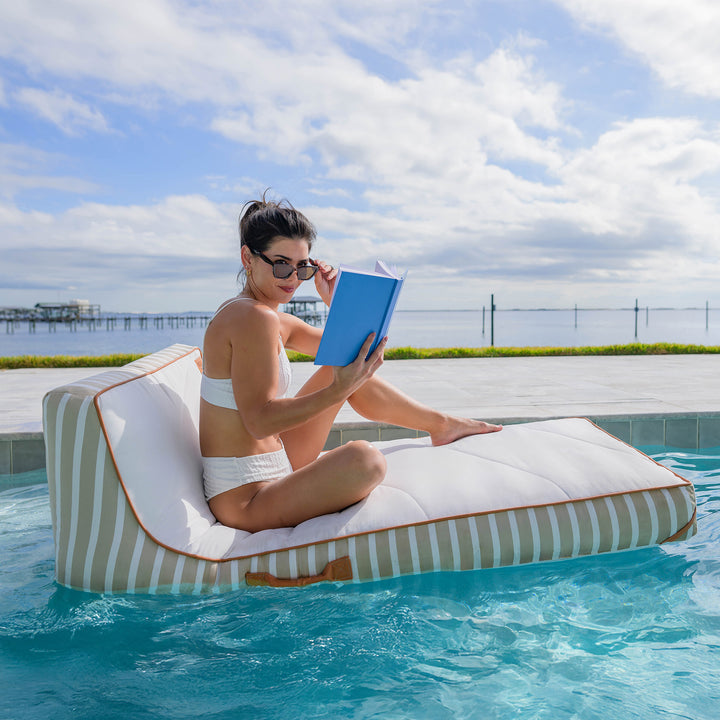 Woman reading a book on a striped pool float in a pool with a scenic background #color_sand-stripe-sunbrella-fabric