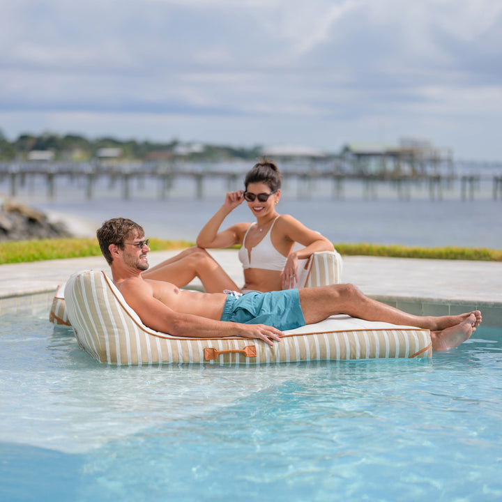 Man and woman relaxing on a striped inflatable lounger by a pool with a scenic background. #color_sand-stripe-sunbrella-fabric
