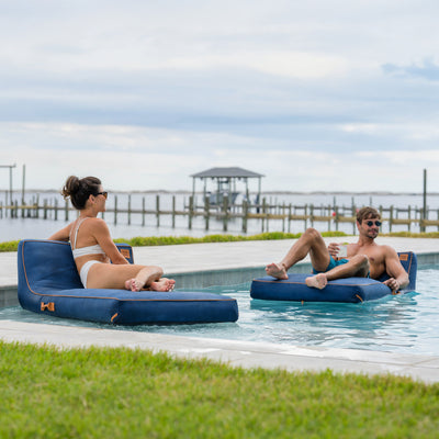 Two people lounging on inflatable pool floats by a pool with a scenic background. 