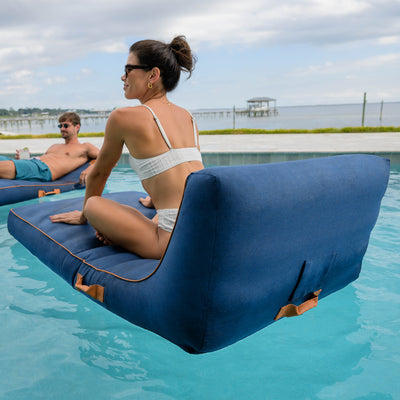 Two people relaxing on inflatable lounge chairs in a pool with a scenic background. 