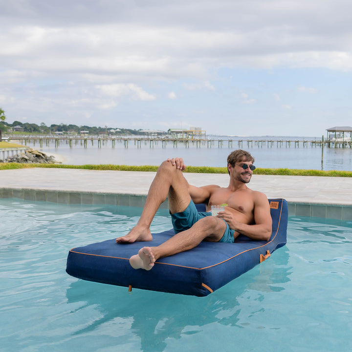 Man lounging on a blue inflatable pool float by a pool with a scenic background #color_harbor-blue-sunbrella-fabric
