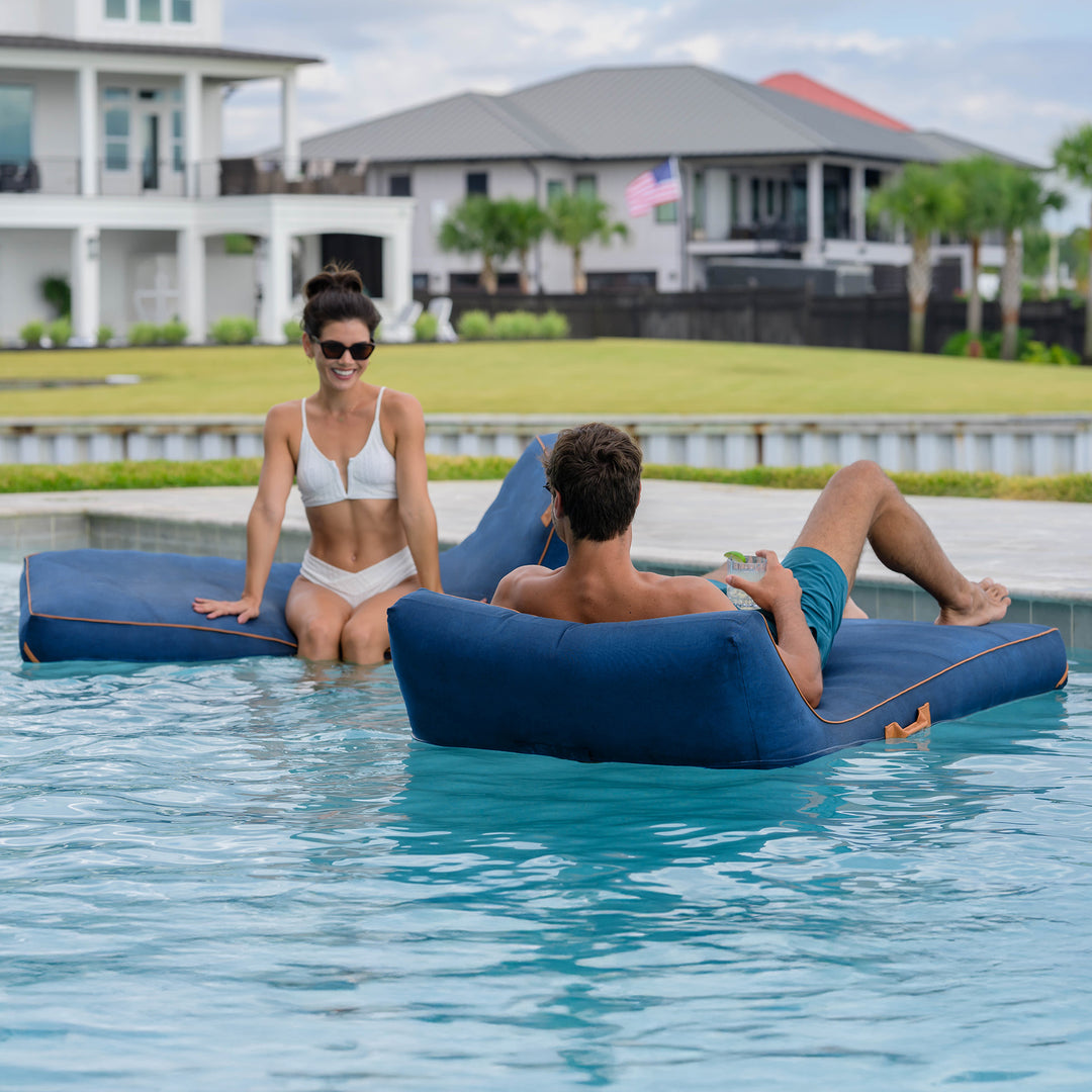 Two people lounging on inflatable blue lounge chairs in a pool with houses and greenery in the background. #color_harbor-blue-sunbrella-fabric