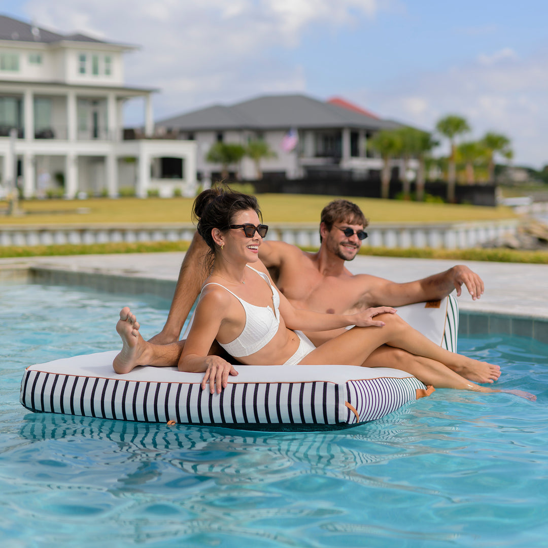 Two people sitting on a striped pool float in a swimming pool with houses and greenery in the background.