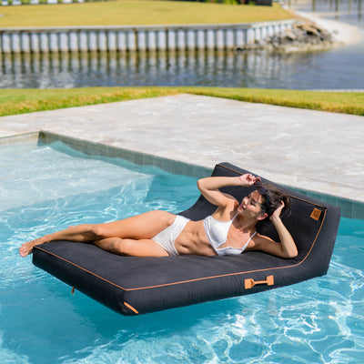 Woman lying on a floating pool mat by a poolside with grass and water in the background 