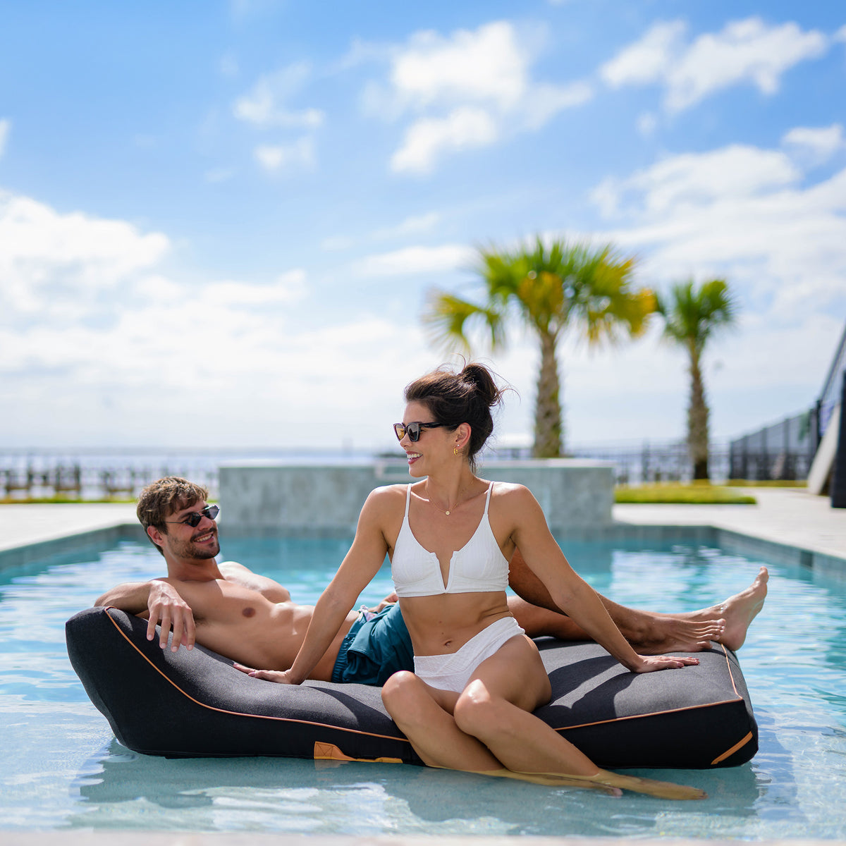 Man and woman relaxing on a pool float in a pool with palm trees in the background 