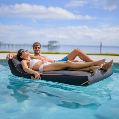 Two people relaxing on a floating platform in a pool with a scenic background. 