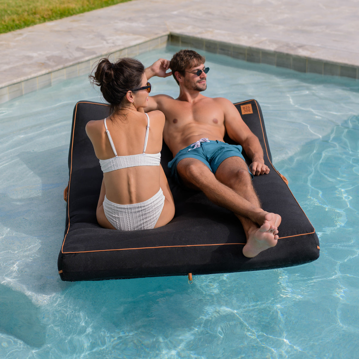 Two people relaxing on a black inflatable pool float in a pool. 