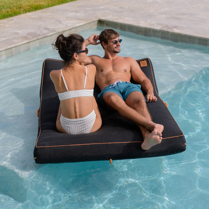 Two people relaxing on a black inflatable pool float in a pool. #color_twilight-sunbrella-fabric