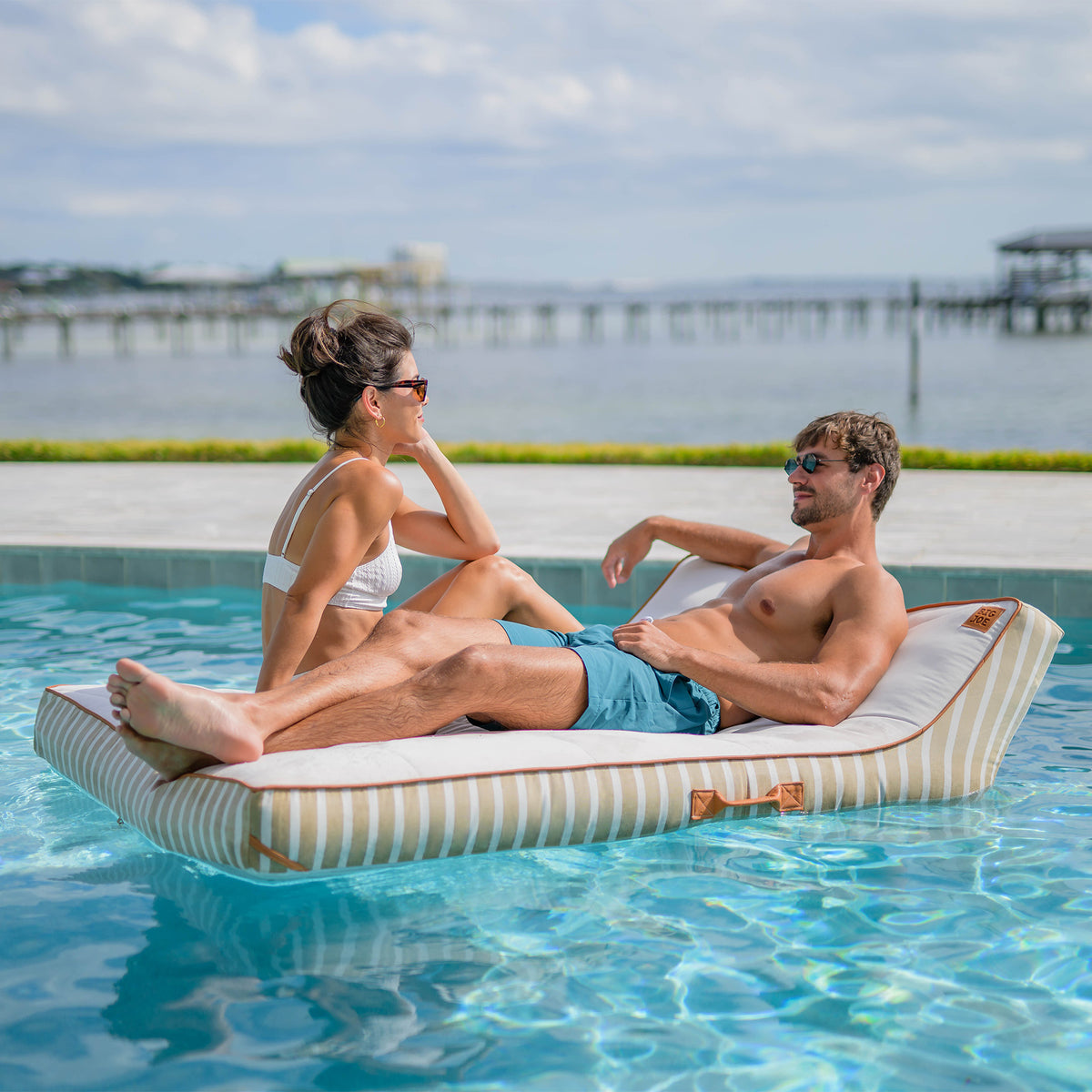 Two people relaxing on a striped inflatable raft in a pool with a scenic background. 
