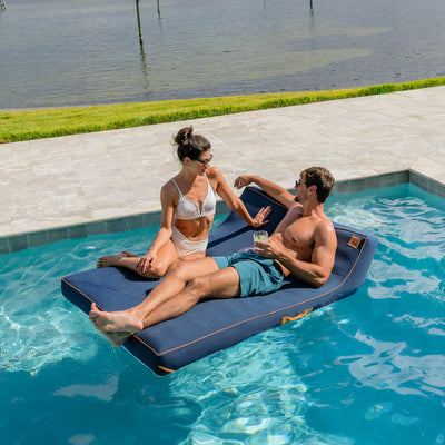 Two people relaxing on a blue inflatable pool float in a swimming pool. 