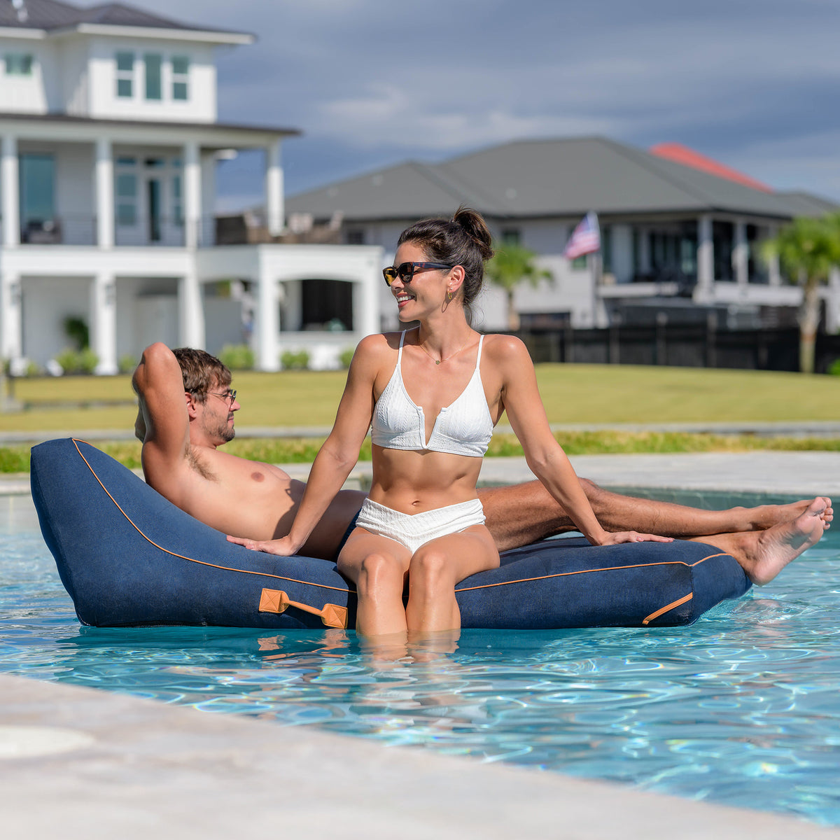 Two people relaxing on a blue inflatable pool float in a swimming pool with houses and greenery in the background. 