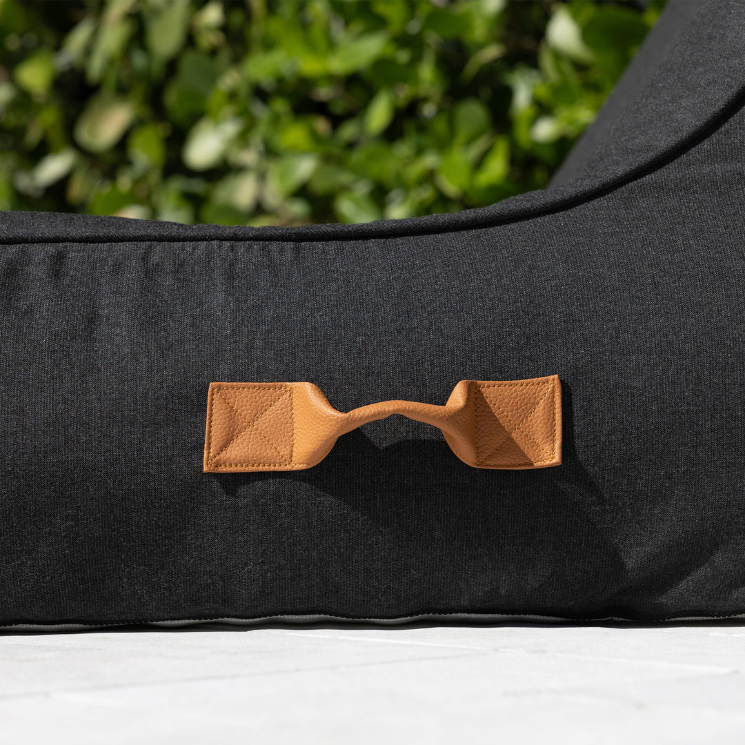 Black pool float lounger with a brown leather handle against a green outdoor background #color_twilight-sunbrella-fabric