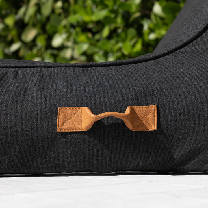 Black pool float lounger with a brown leather handle against a green outdoor background #color_twilight-sunbrella-fabric