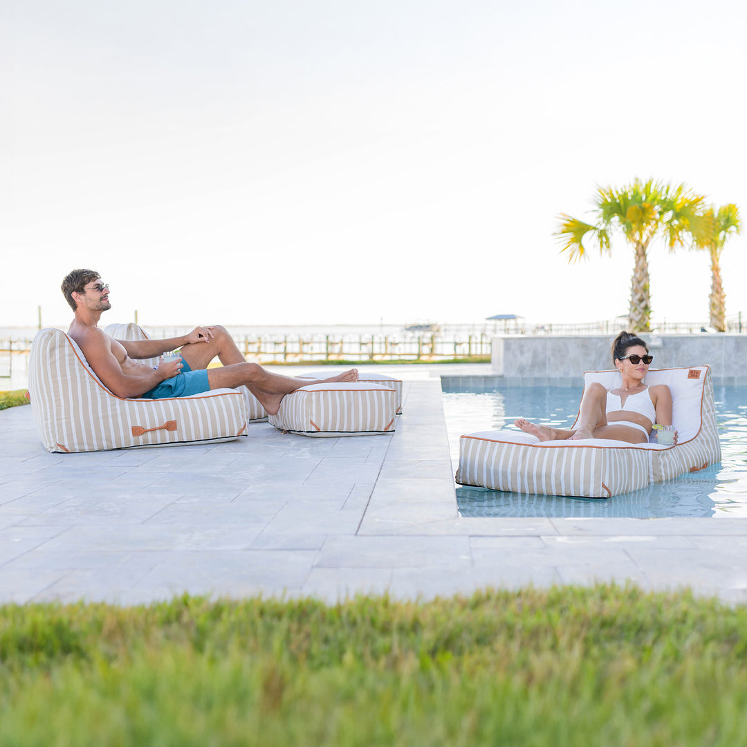 Two people relaxing on striped bean bags by a pool with palm trees in the background. #color_sand-stripe-sunbrella-fabric