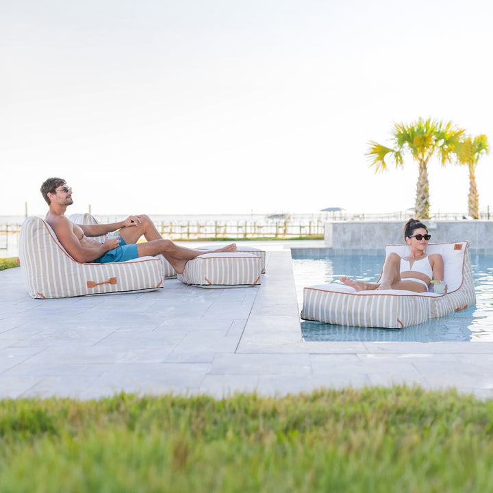 Two people relaxing on striped bean bags by a pool with palm trees in the background. #color_sand-stripe-sunbrella-fabric