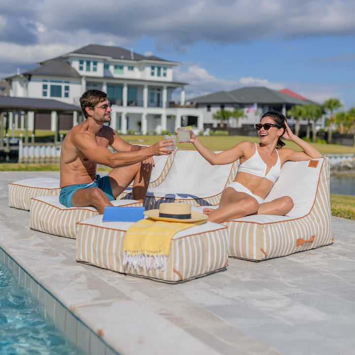 Two people sitting on lounge chairs by a pool with a house and palm trees in the background. #color_sand-stripe-sunbrella-fabric