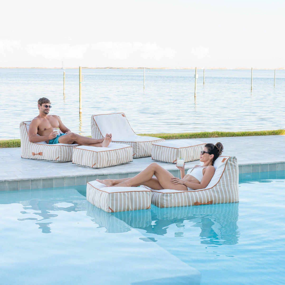 Two people relaxing on striped lounge chairs by a pool with a scenic background. #color_sand-stripe-sunbrella-fabric
