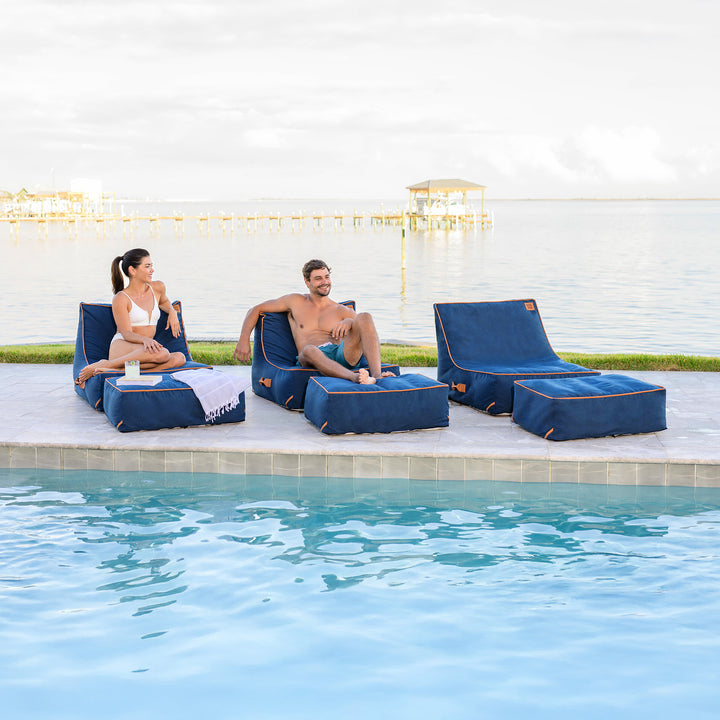 Two people sitting on blue lounge chairs by a poolside. #color_harbor-blue-sunbrella-fabric