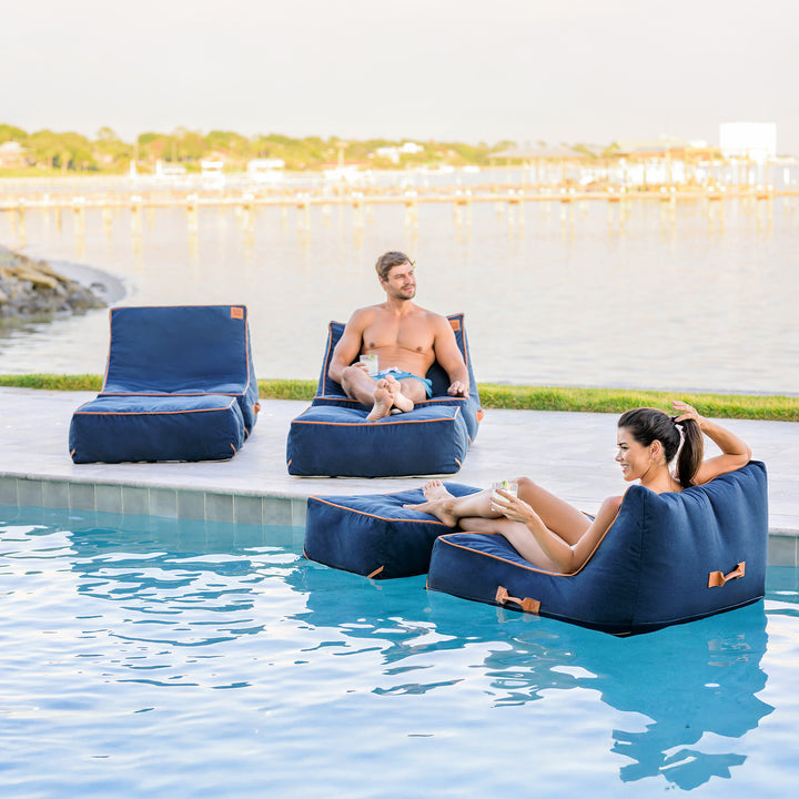 Two people relaxing on blue lounge chairs by a pool. #color_harbor-blue-sunbrella-fabric