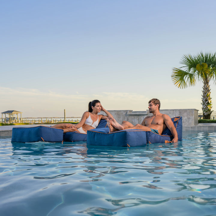 Two people relaxing on blue lounge chairs in a pool with palm trees in the background. #color_harbor-blue-sunbrella-fabric