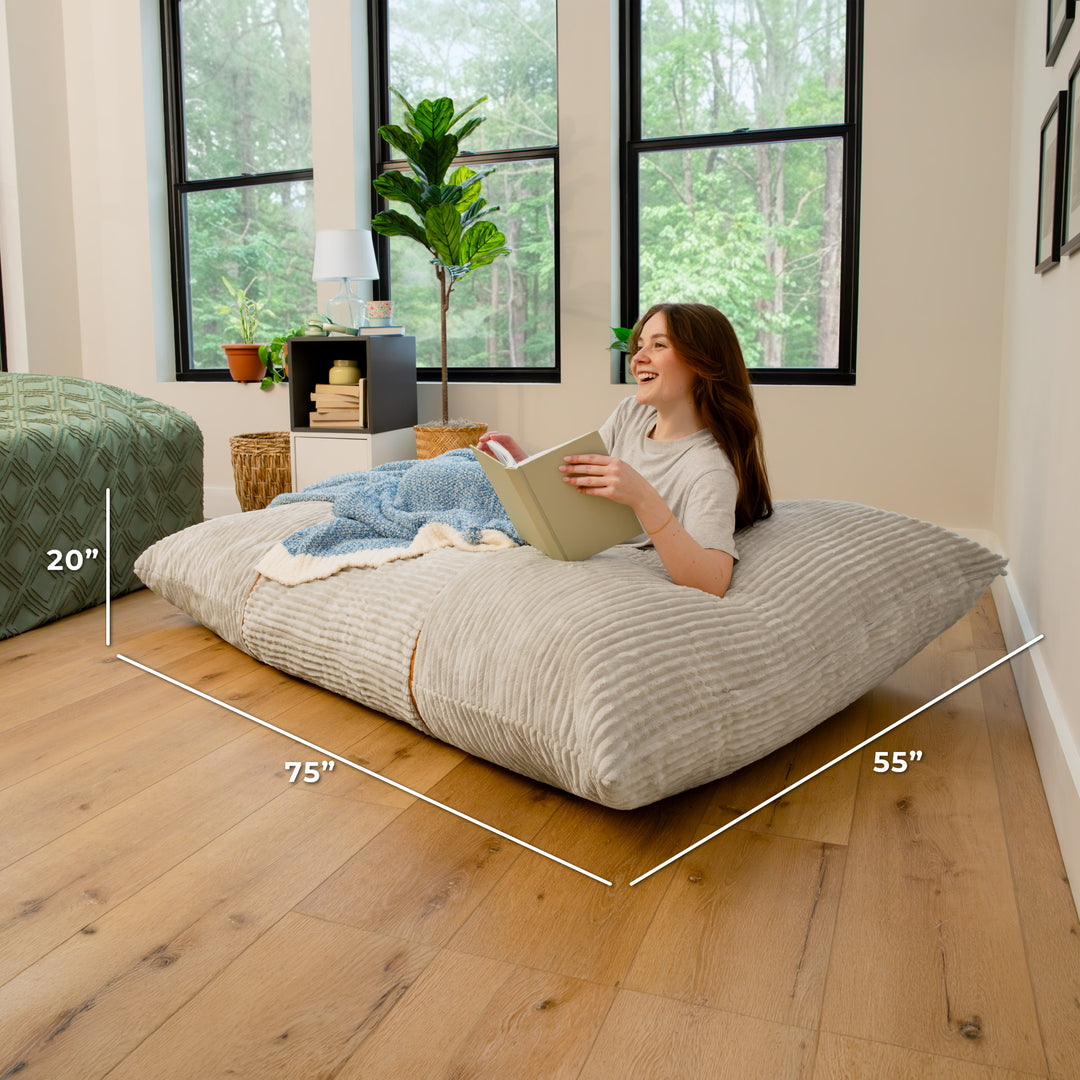 A woman sits on a large floor cushion in a bright room with wood floors and large windows. White dimension lines and text show the cushion measurements as 75 inches by 55 inches by 20 inches. 