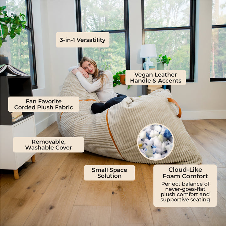 A woman reclines on a large beige corded plush bean bag chair in a bright room with wood floors and large windows. Text callouts describe features such as 3-in-1 versatility, vegan leather handle and accents, removable washable cover, small space solution, and cloud-like foam comfort. 