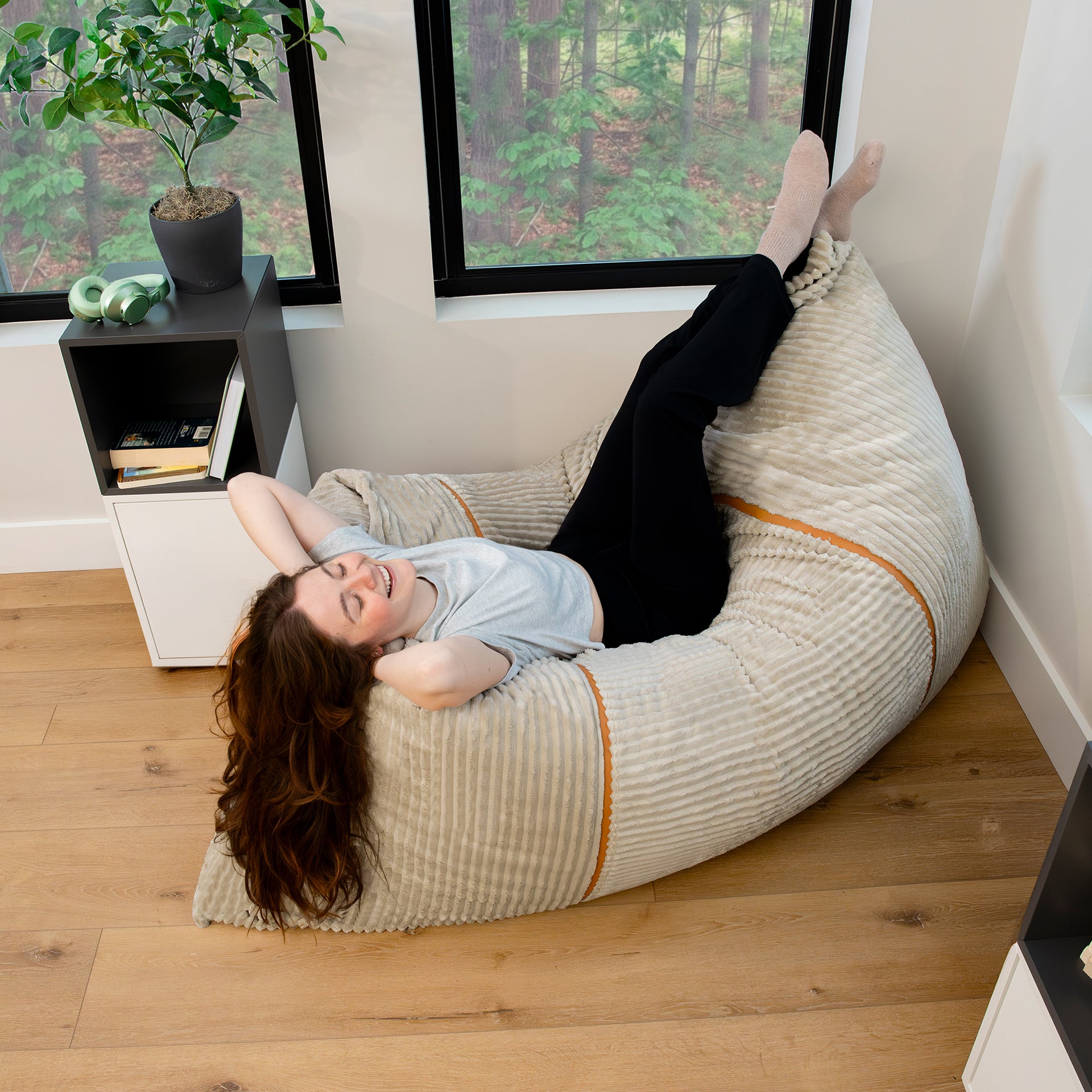 A woman reclining on a large striped beige bean bag chair in a modern room with wood floors and windows looking out to a forest. #color_vanilla-latte-corded-plush