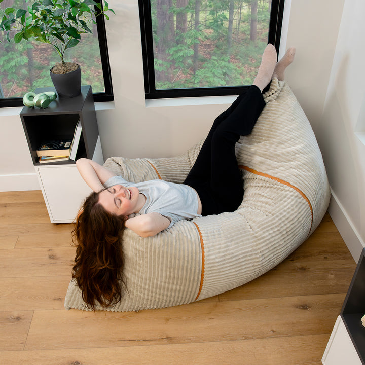 A woman reclining on a large striped beige bean bag chair in a modern room with wood floors and windows looking out to a forest. 