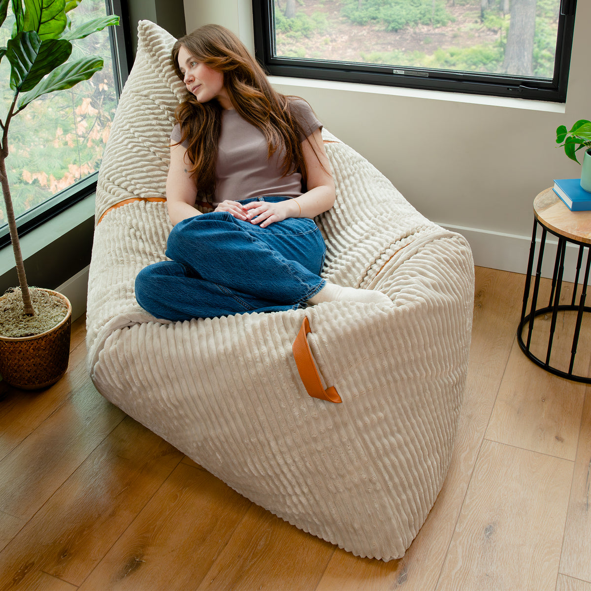 A woman reclines in a large cream ribbed bean bag chair with brown leather handles in a bright room with wood floors, a window, and indoor plants. #color_vanilla-latte-corded-plush