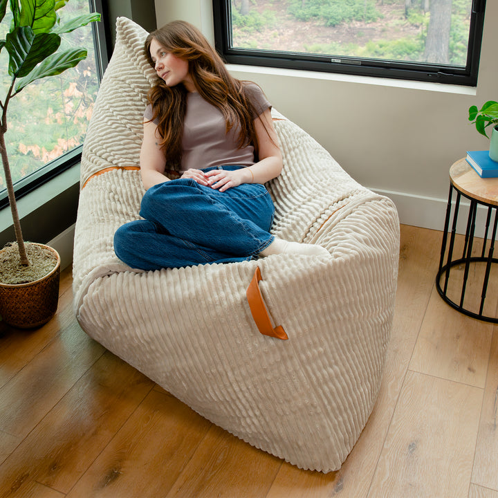 A woman reclines in a large cream ribbed bean bag chair with brown leather handles in a bright room with wood floors, a window, and indoor plants. 
