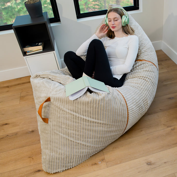 A woman relaxes in a large cream ribbed bean bag chair with orange-brown handles, sitting in a bright room with wood floors and a small side table. 
