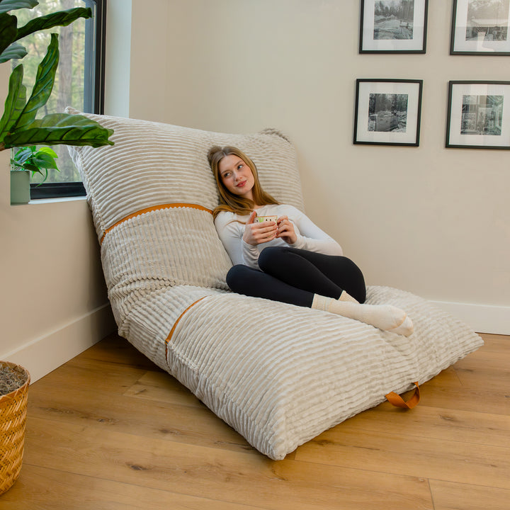 A woman sits on a large, ribbed, floor-launched lounger in a cozy room corner with light wood floors, a potted plant, and framed wall art. 