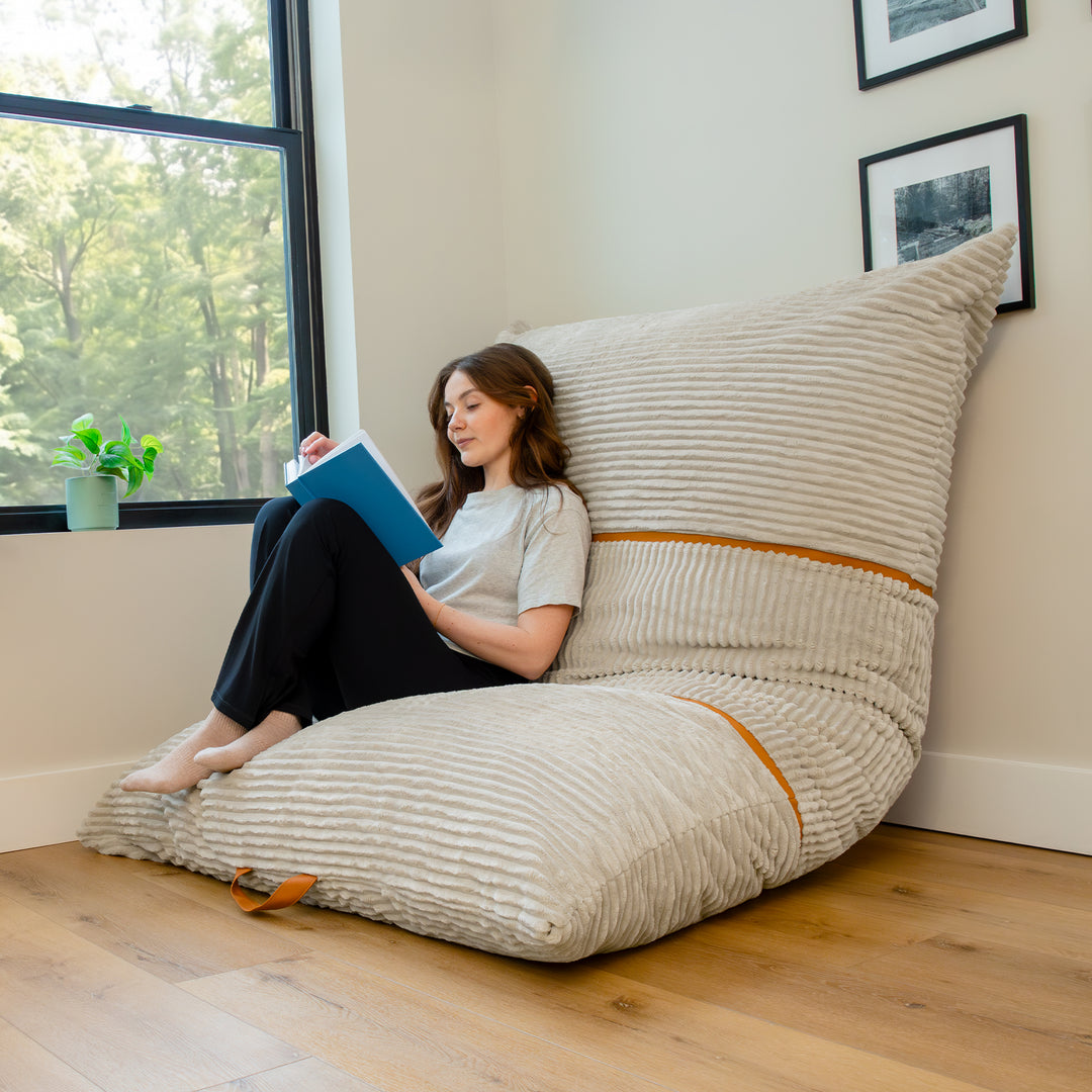 A woman sits on a large ribbed wedge floor cushion in a bright room, reading a blue book. The cushion is upholstered in a light beige textured fabric with subtle orange-brown accent stripes and is positioned against a wall near a window. 
